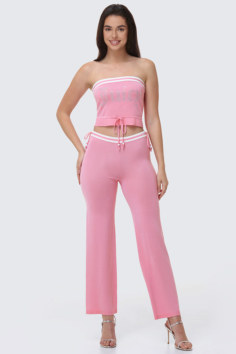 A woman smiles in the Big Bling Tube Top, a pink strapless crop top with white trim and decorative text, paired with matching flared pants and clear high-heeled sandals against a light gray background.