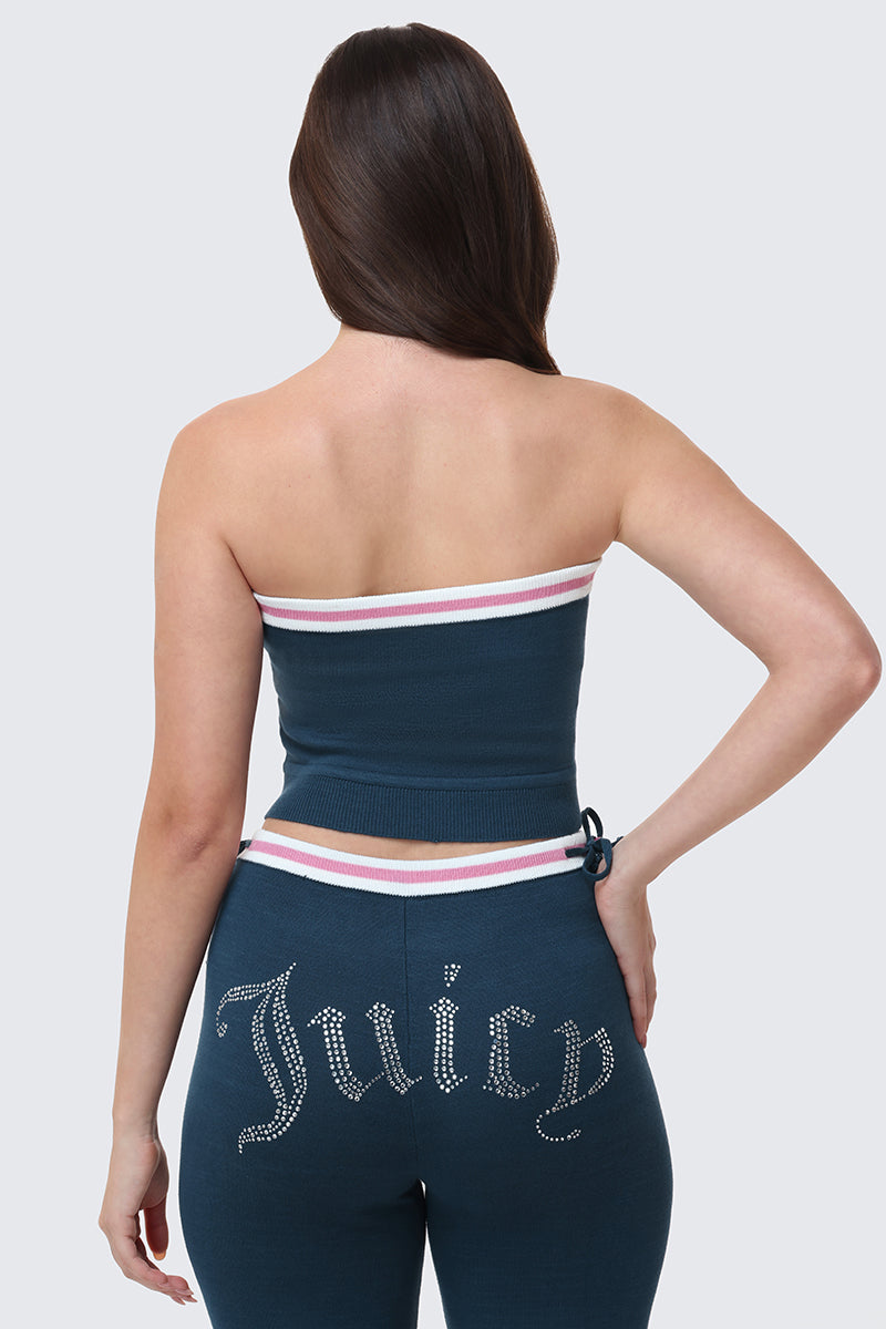 A woman with long brown hair faces away, wearing the Big Bling Tube Top—a dark blue strapless top with white and pink stripes—and matching pants featuring "Juicy" in rhinestones on the back.
