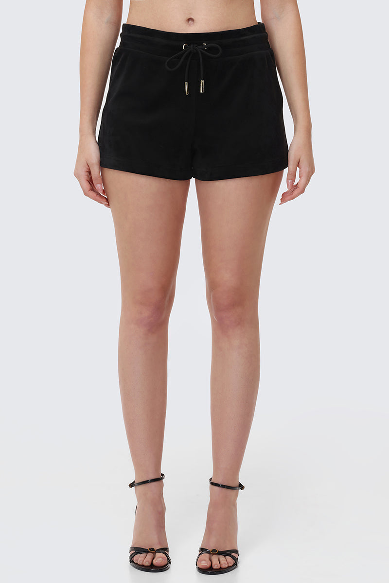 A woman stands facing forward in Big Bling Velour Shorts with a drawstring waist and black strappy high-heeled sandals. Her arms are relaxed at her sides against a plain light gray background.