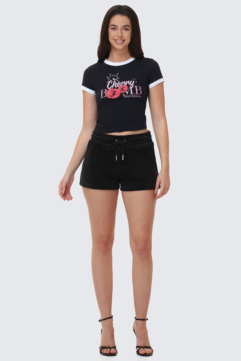 A woman faces forward, smiling, wearing a black Cherry Babe graphic crop top, Big Bling Velour Shorts with a drawstring waist, and black high-heeled sandals against a light gray background.
