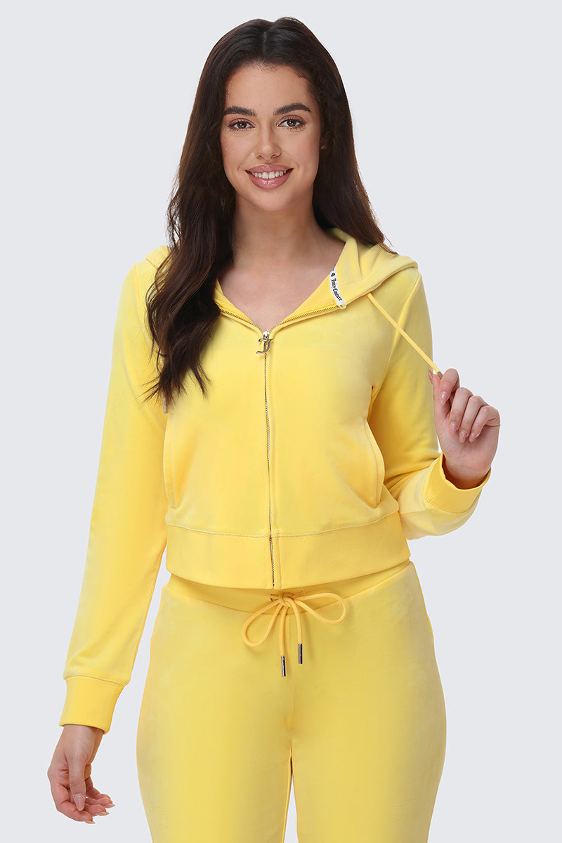 A woman with long dark hair smiles while wearing the OG Big Bling Velour Hoodie in yellow and matching drawstring sweatpants, standing against a plain light background.