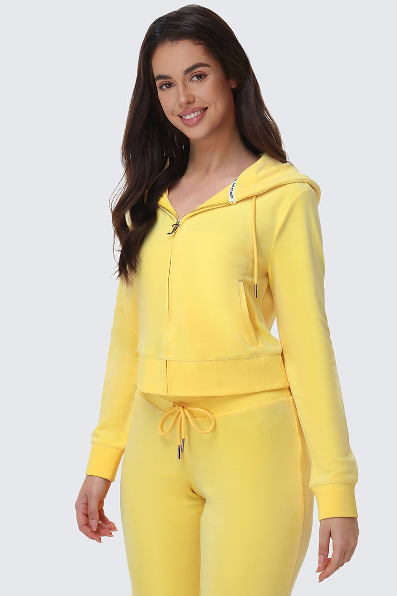 A woman with long dark hair is smiling and wearing the OG Big Bling Velour Hoodie with matching yellow sweatpants, standing against a light gray background.