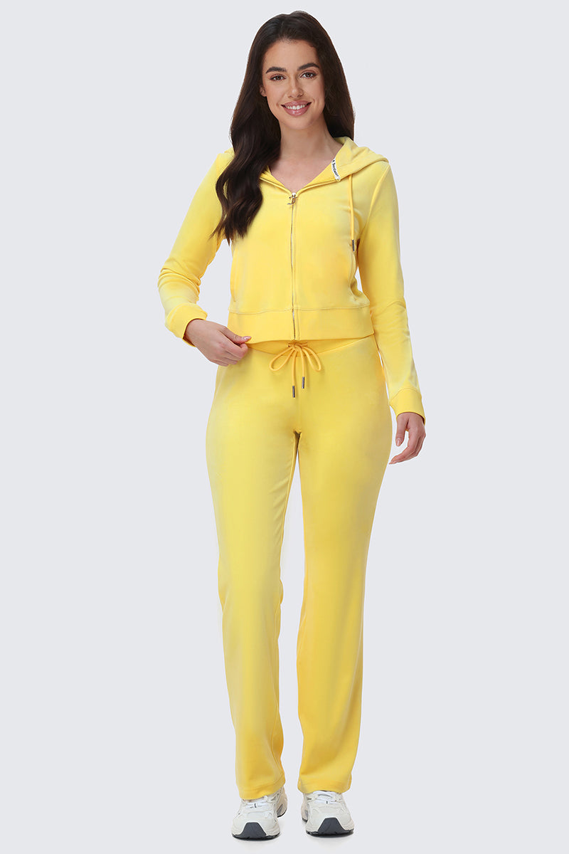 A woman smiles wearing the OG Big Bling Velour Hoodie in yellow with matching drawstring sweatpants and white sneakers, posed against a plain light gray background.