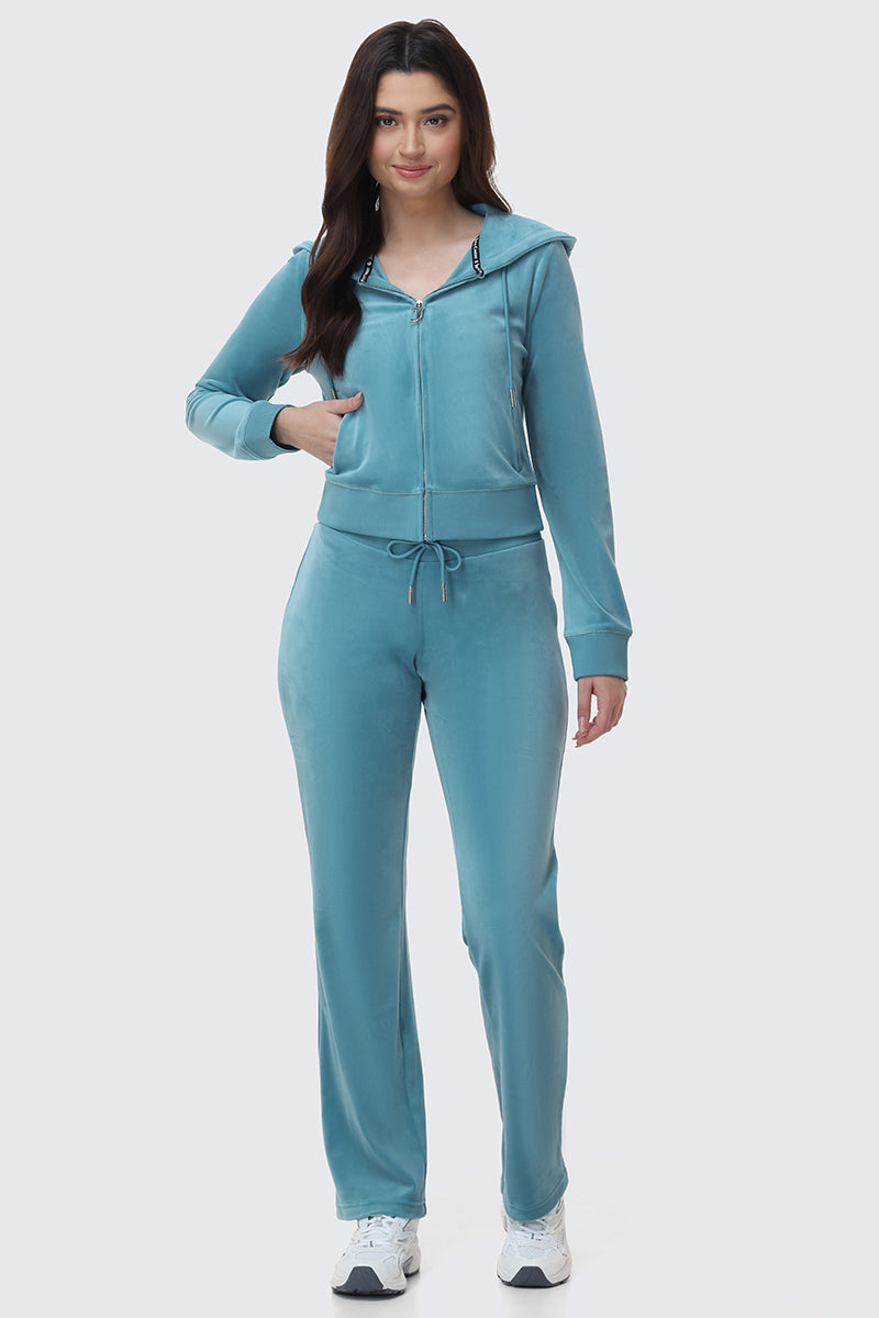 A woman faces forward, wearing the OG Big Bling Velour Hoodie in light blue with matching drawstring pants and white sneakers. She smiles softly, one hand in her pocket, against a plain light background.