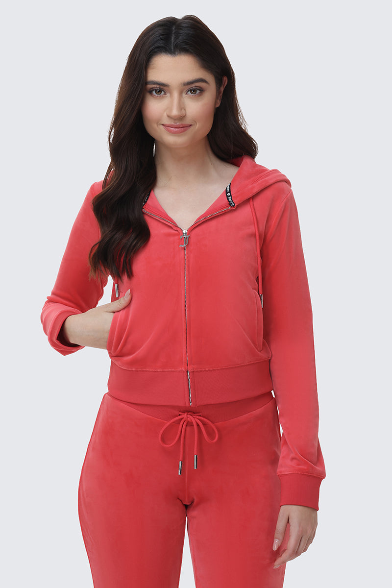 A woman with long dark hair models the OG Big Bling Velour Hoodie and matching drawstring pants in red, standing against a light gray background and smiling softly at the camera.
