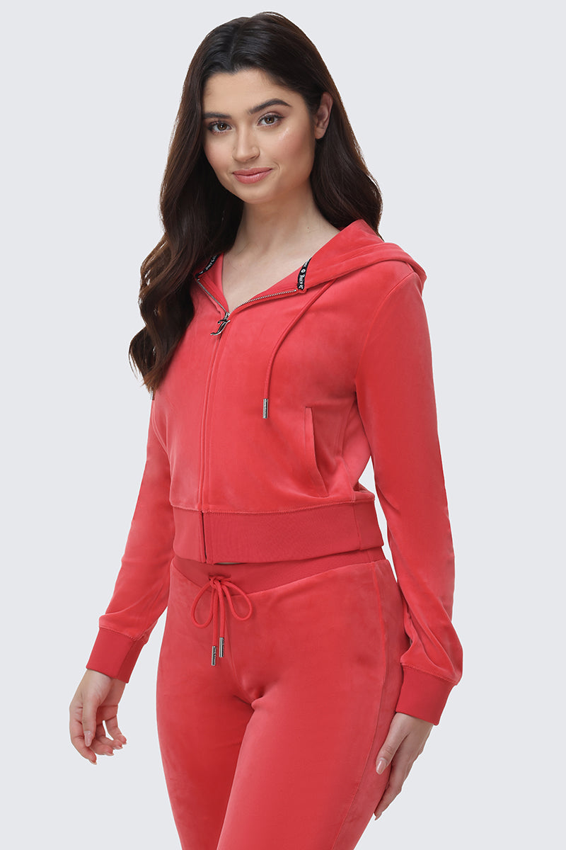 A woman with long dark hair smiles gently in the OG Big Bling Velour Hoodie and matching drawstring pants, standing against a plain light background—a luxe update to classic comfort.