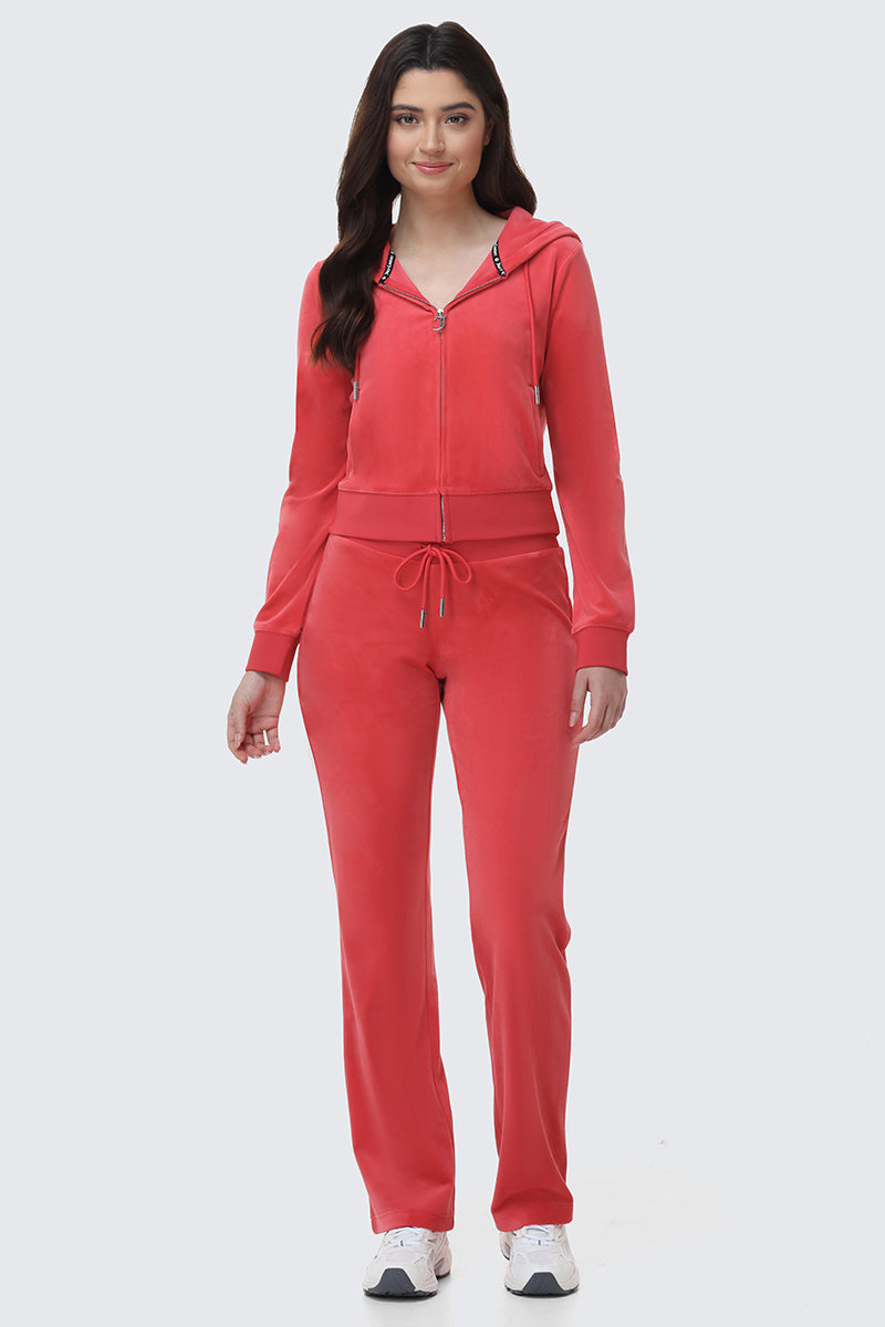A woman faces forward in the OG Big Bling Velour Hoodie and matching drawstring pants, both in coral pink. Her long dark hair and white sneakers complete this luxe take on classic loungewear, set against a plain light background.