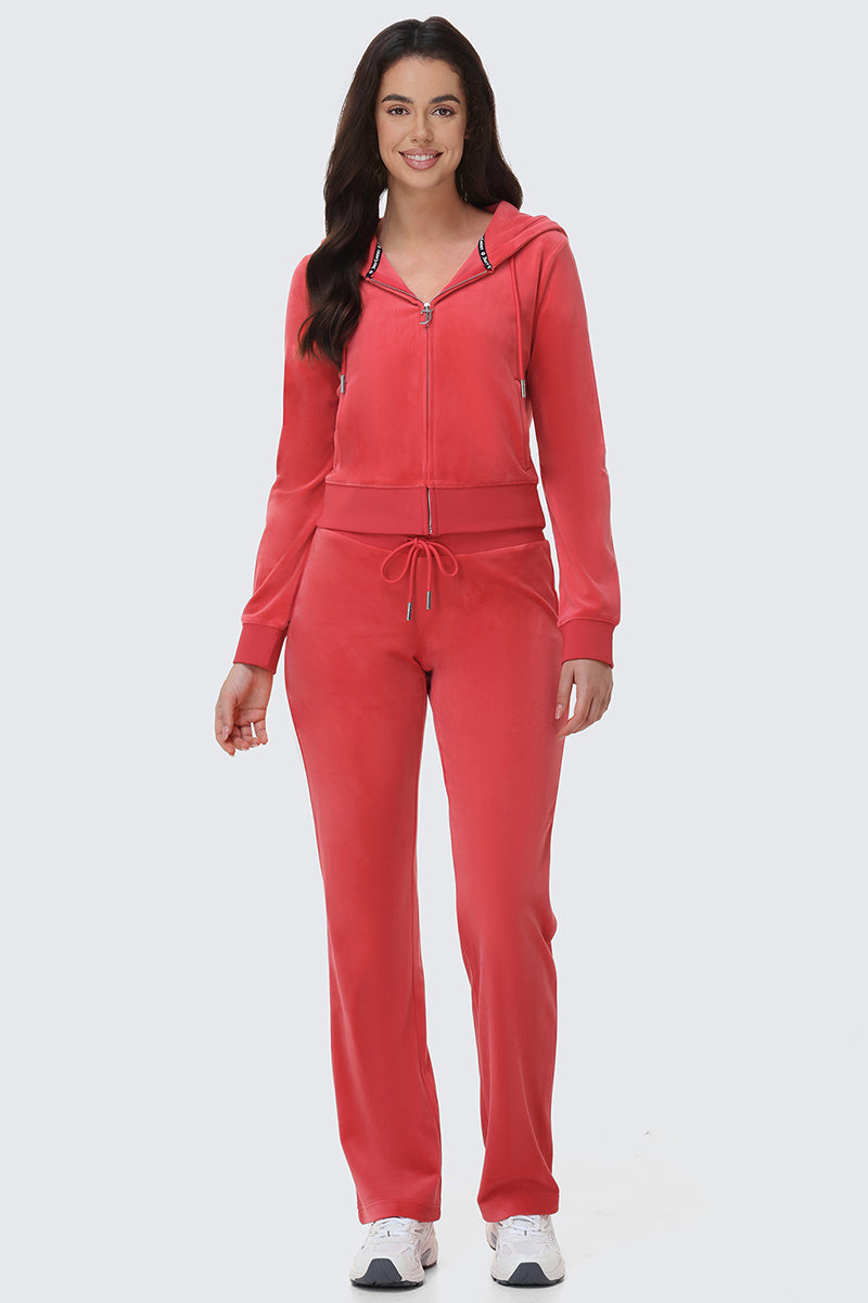 A woman smiles in the OG Big Bling Velour Hoodie, a coral pink tracksuit with drawstring waist pants and white sneakers, standing against a light grey background.