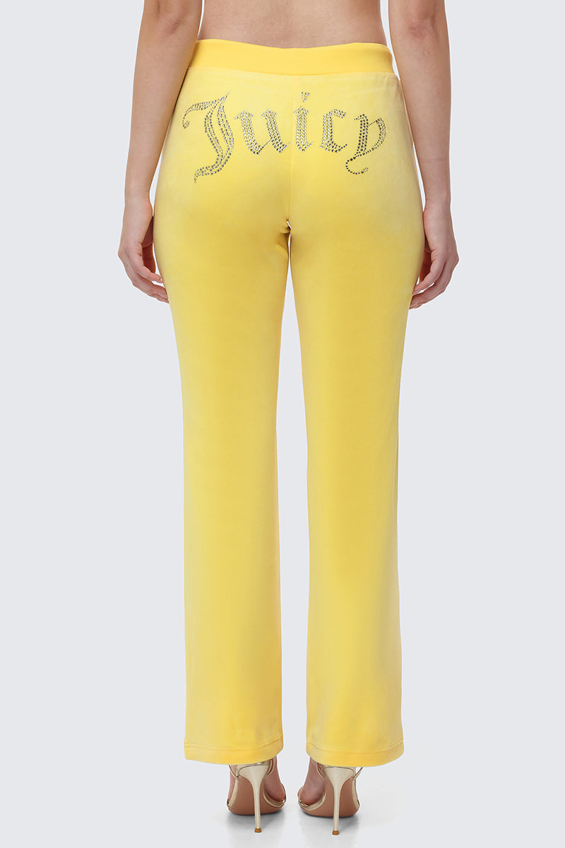 Someone models the OG Big Bling Velour Track Pants—yellow, Y2K-inspired velour pants with rhinestone "Juicy" embellishment on the back—standing against a plain light background.