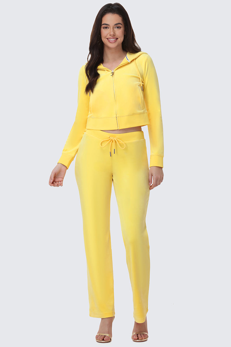 A woman smiles in OG Big Bling Velour Track Pants—Y2K-inspired, bright yellow, with a matching zip-up hoodie and drawstring—paired with open-toed heels against a plain light gray background.