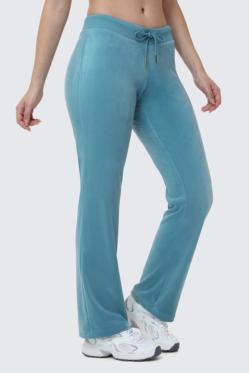 Wearing OG Big Bling Velour Track Pants in teal with white sneakers, a person poses with one leg slightly bent against a light gray background, capturing a cool Y2K vibe.