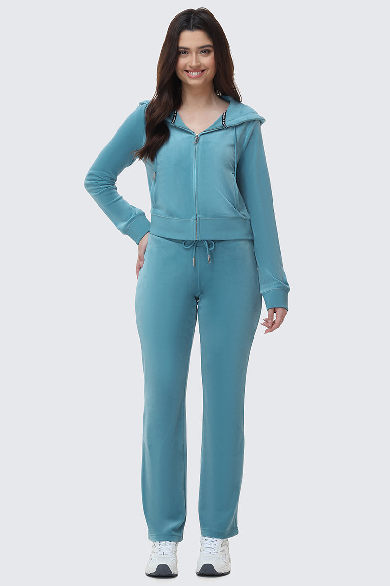 A woman smiles while wearing OG Big Bling Velour Track Pants and a light blue zip-up hoodie with a drawstring waist, paired with white sneakers, standing against a plain light background.