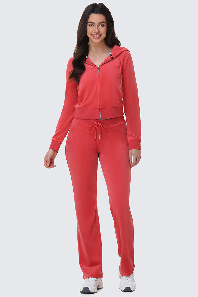 A woman with long dark hair wears OG Big Bling Velour Track Pants and a matching red zip-up hoodie featuring Juicy embellishment. She smiles in white sneakers against a plain light gray background.
