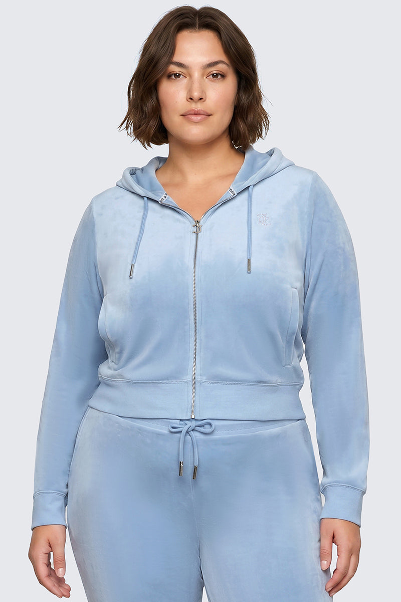 A woman wearing the Plus-Size OG Big Bling Velour Hoodie in light blue, featuring a subtle Juicy logo and matching drawstring pants, stands facing the camera and channels effortless Y2K style against a neutral background, her expression calm and composed.