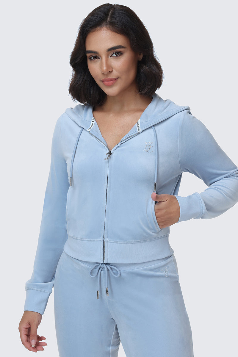 A woman with dark, shoulder-length hair wears the OG Big Bling Velour Hoodie—a light blue zip-up adorned with a rhinestone Juicy logo—along with matching pants, standing against a plain light background with one hand on her hip and a slight smile.