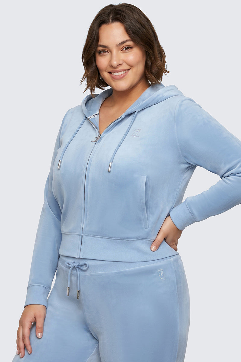 A woman with short brown hair smiles, wearing the Plus-Size OG Big Bling Velour Hoodie in light blue with matching pants, her hand on her hip as she stands against a plain light background.