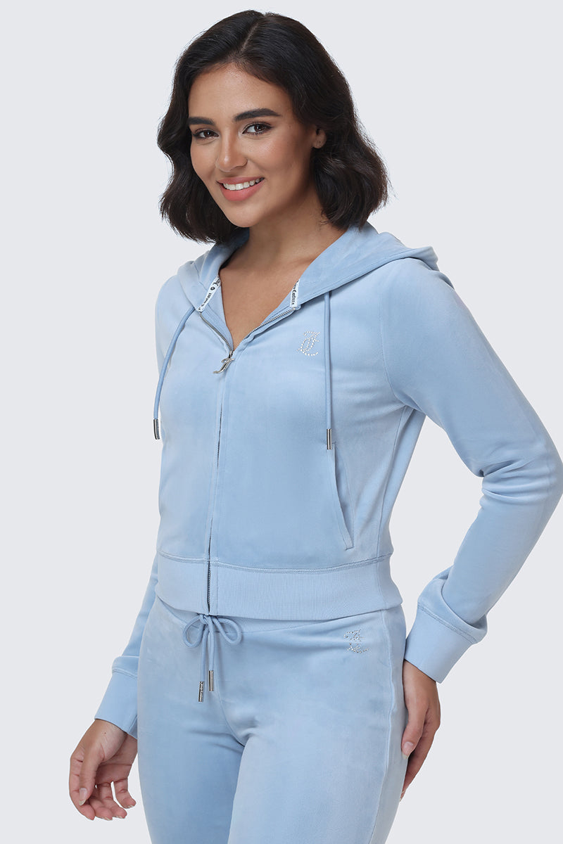 A woman with short dark hair wears the OG Big Bling Velour Hoodie in light blue, featuring a dazzling rhinestone Juicy logo on the back, smiling and standing against a plain light background—capturing iconic Y2K Juicy style.