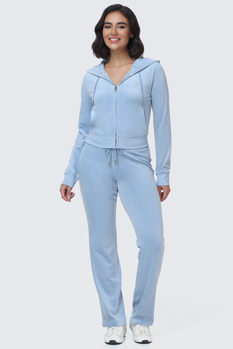 A woman stands smiling on a plain light background, wearing the OG Big Bling Velour Hoodie in light blue—a zip-up, bedazzled velour hoodie—paired with matching drawstring pants and white sneakers.
