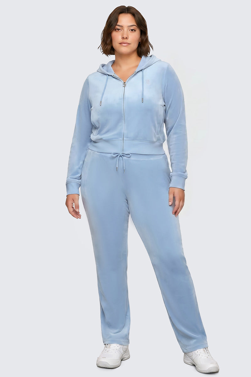 A woman stands facing forward, wearing the Plus-Size OG Big Bling Velour Hoodie in light blue with a rhinestone Juicy logo, paired with matching drawstring pants and white sneakers, against a plain light background.