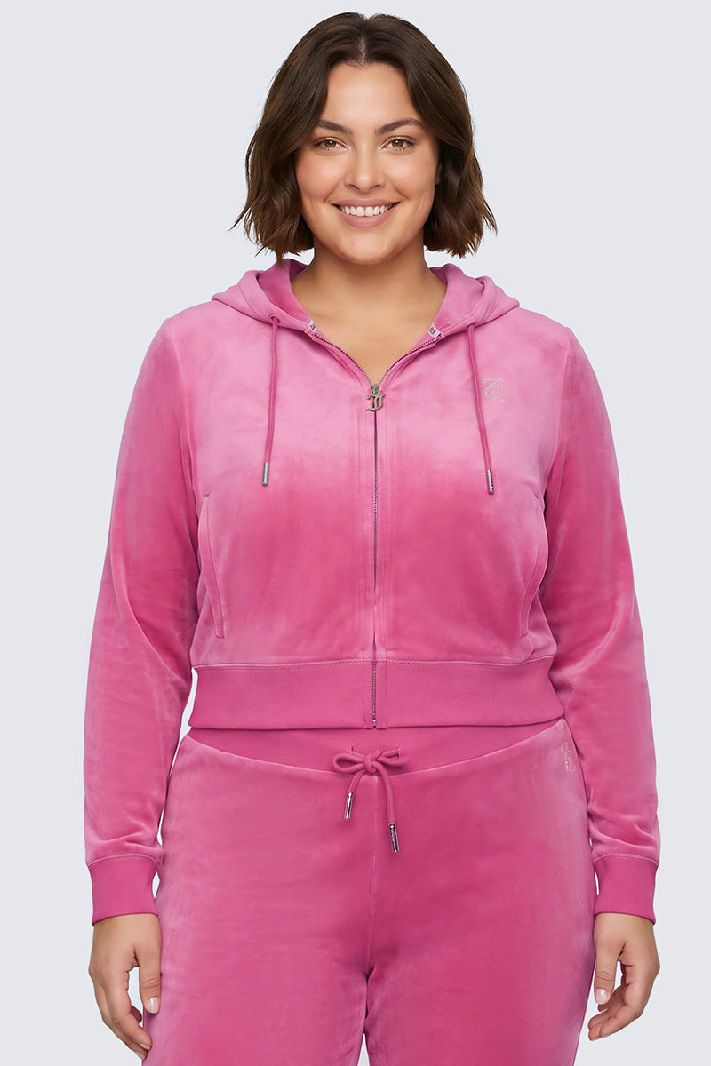 A woman smiles while wearing the Plus-Size OG Big Bling Velour Hoodie, a zip-up pink velour piece with drawstrings, long sleeves, and a sparkling bedazzled Juicy logo, paired with matching sweatpants for ultimate Y2K vibes. She stands against a plain light background.