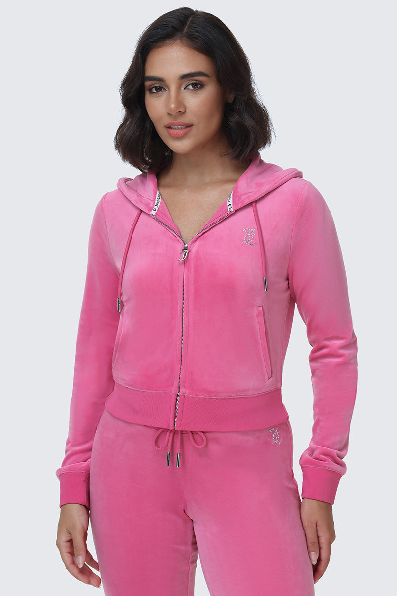 A woman stands against a plain light background, looking at the camera with a slight smile, wearing the OG Big Bling Velour Hoodie and matching pants in pink. The Y2K-inspired zip-up hoodie features sparkling rhinestone detailing on the Juicy logo, adding playful glamour to her outfit.