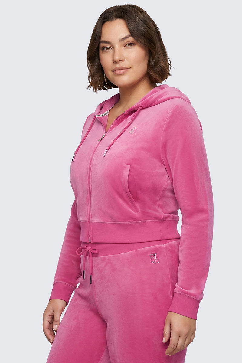 A woman with short brown hair stands confidently against a plain light gray background, embodying true Y2K style as she wears the Plus-Size OG Big Bling Velour Hoodie in pink, which features a sparkling bedazzled Juicy logo and matching sweatpants.