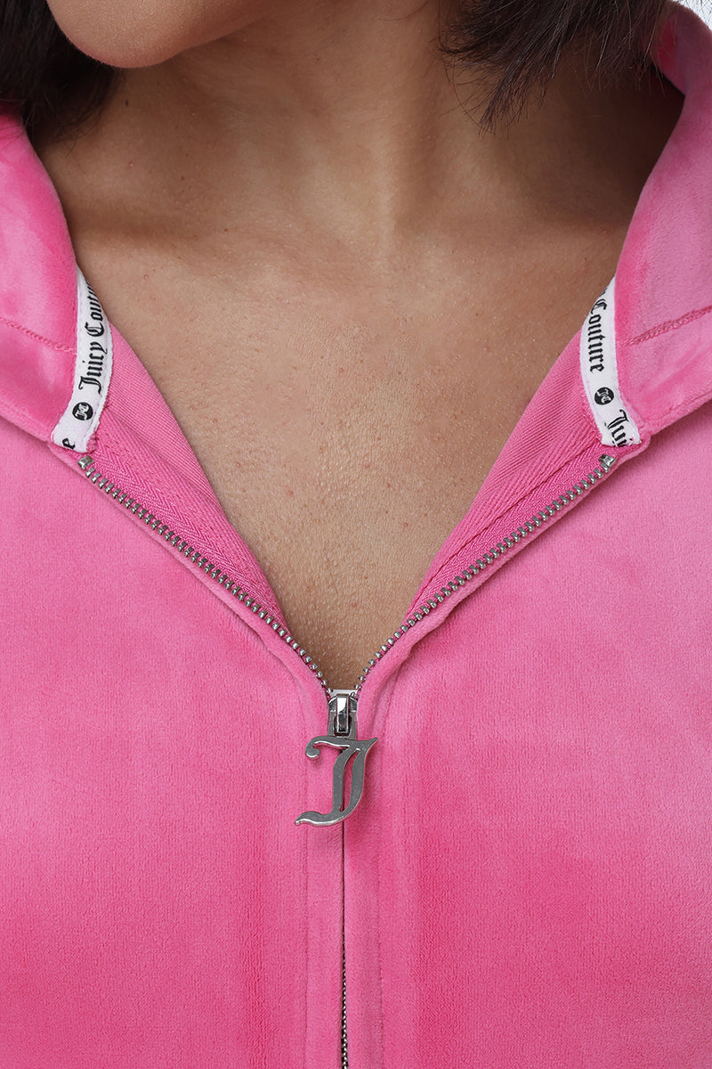 Close-up of a person wearing the OG Big Bling Velour Hoodie in pink, featuring a silver J zipper pull and white trim with “Juicy Couture” lettering. The slightly unzipped hoodie reveals part of the neck and collarbone, capturing classic Y2K style.