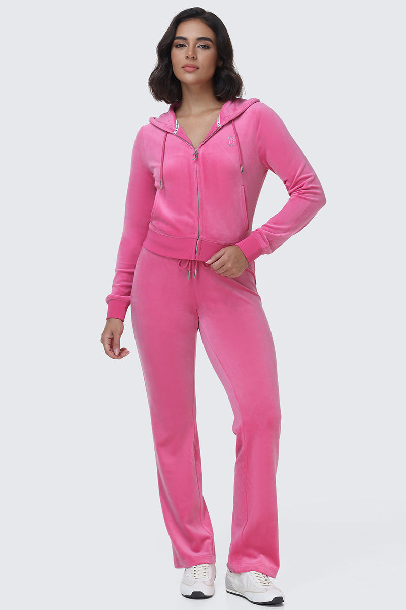 A woman stands against a plain light gray background in the perfect Y2K-inspired look, wearing the OG Big Bling Velour Hoodie—a bright pink velour zip-up with a sparkling rhinestone Juicy logo—paired with matching drawstring pants and crisp white sneakers.