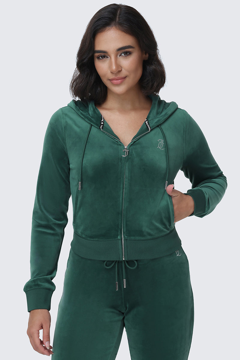A woman with shoulder-length dark hair wears the OG Big Bling Velour Hoodie—a green, zip-up velour tracksuit top adorned with rhinestones—paired with matching pants featuring a sparkling Juicy logo, standing against a plain light gray background.