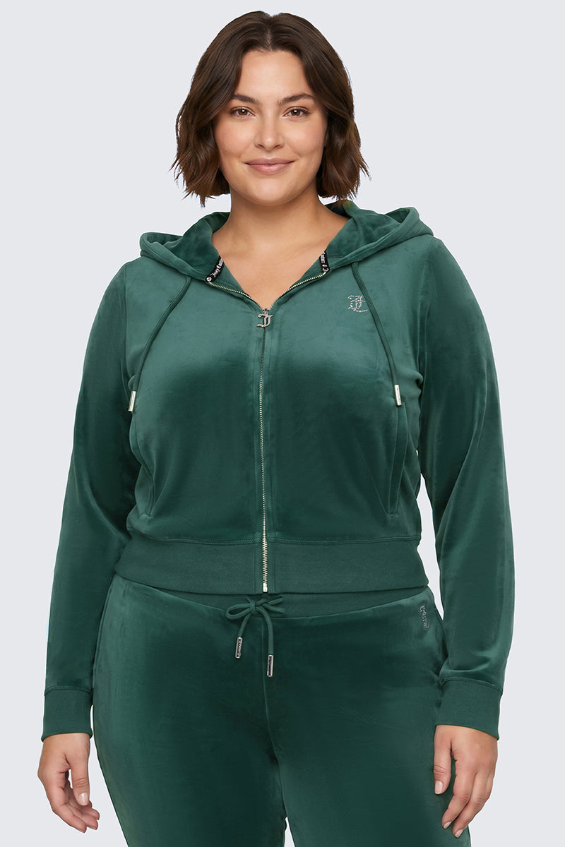 A person stands smiling against a plain light gray background, wearing the Plus-Size OG Big Bling Velour Hoodie in dark green with a sparkling rhinestone Juicy logo, paired with matching pants.