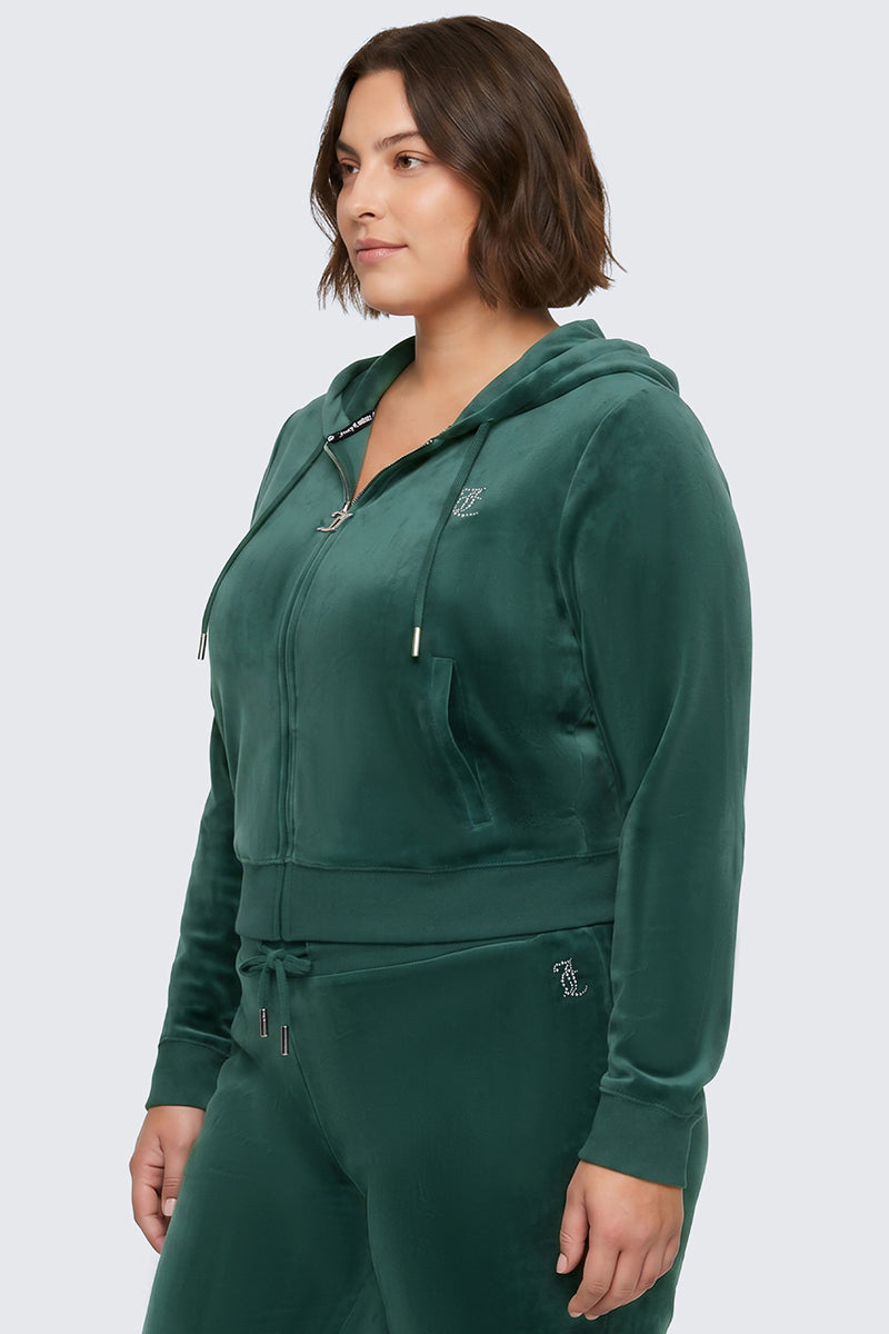 A woman is wearing the Plus-Size OG Big Bling Velour Hoodie in forest green—a zip-up, bedazzled hoodie paired with drawstring pants featuring a rhinestone Juicy logo—standing against a plain light gray background.