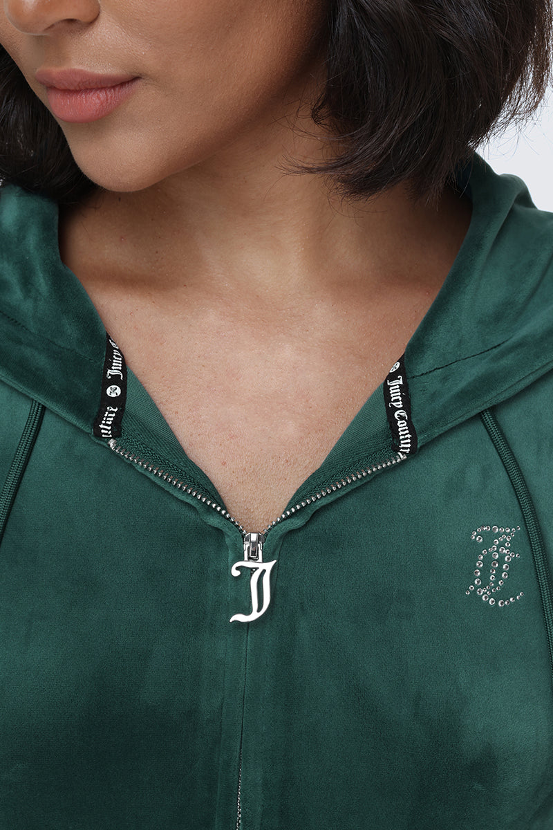 A woman wearing the OG Big Bling Velour Hoodie in green, featuring a sparkling J charm on the zipper and rhinestone Juicy logo across the chest. Only her lower face, neck, and upper chest are visible, evoking that iconic Y2K vibe.