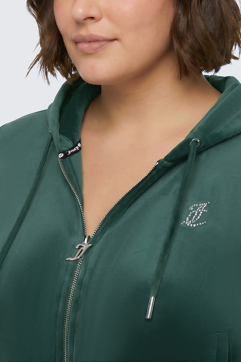 A woman channels effortless Y2K style in the Plus-Size OG Big Bling Velour Hoodie—a dark green zip-up hoodie adorned with a rhinestone Juicy logo on the chest, partially zipped to reveal a glimpse of a black shirt underneath.