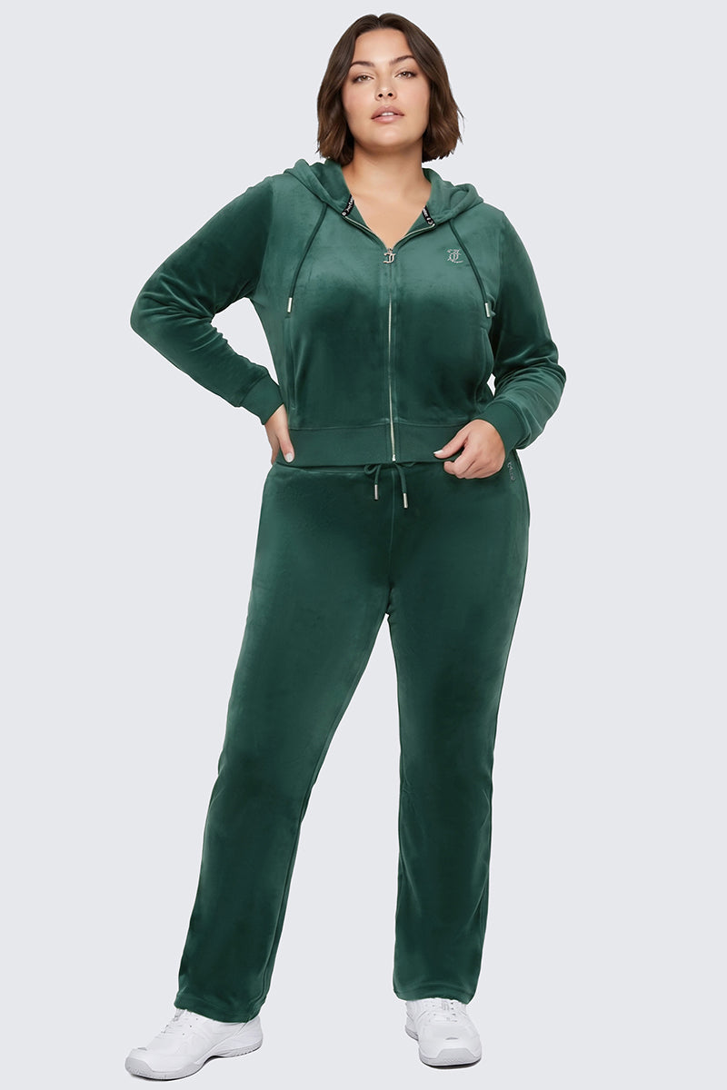 A woman stands with one hand in her pocket, wearing the Plus-Size OG Big Bling Velour Hoodie—a green velour tracksuit with a bedazzled rhinestone Juicy logo on the hoodie and matching pants. She completes her classic Y2K look with white sneakers.