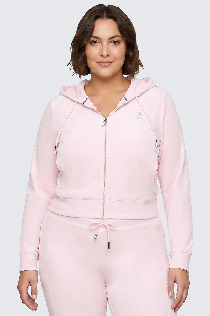 A woman with short brown hair wears the Plus-Size OG Big Bling Velour Hoodie in light pink, featuring a rhinestone Juicy logo, paired with matching drawstring joggers. She stands facing forward, smiling softly with her hands relaxed at her sides against a plain light gray background.