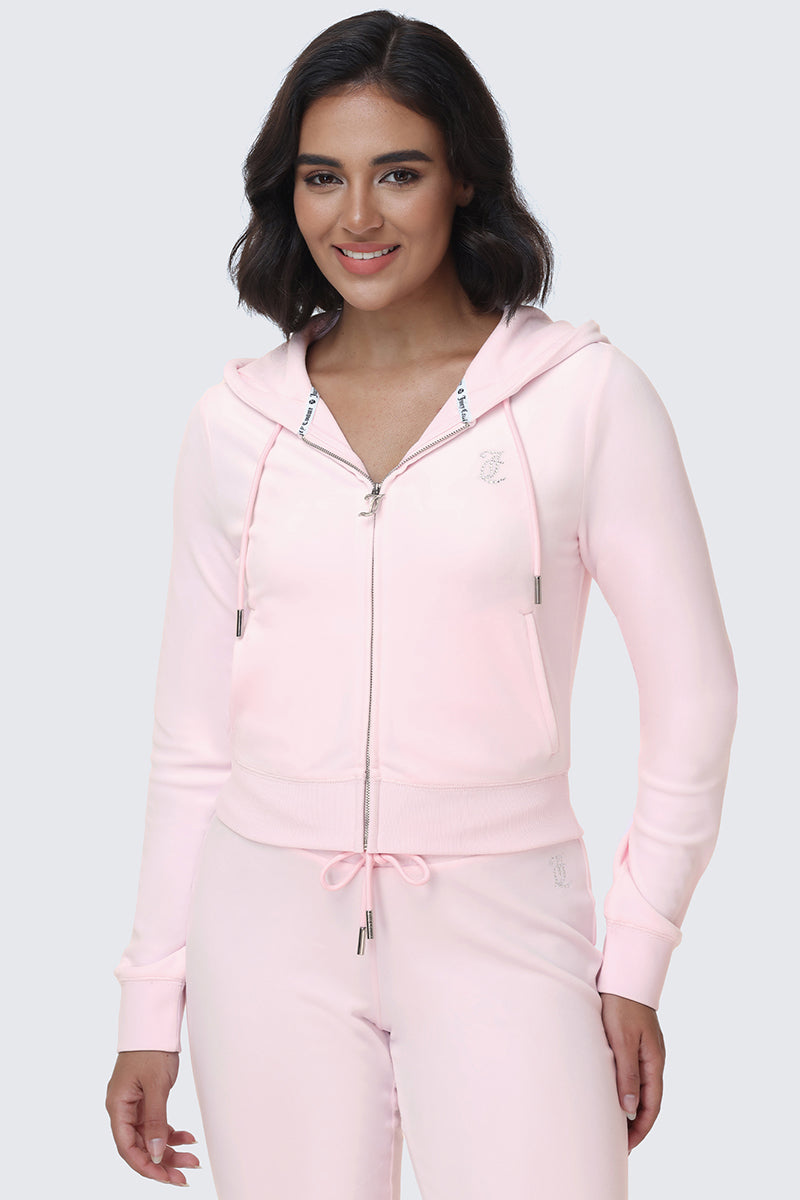 A woman with shoulder-length dark hair smiles while wearing the OG Big Bling Velour Hoodie—a light pink, bedazzled zip-up featuring a drawstring, front pockets, and subtle Y2K vibes—paired with matching pants. She stands against a plain light gray background.