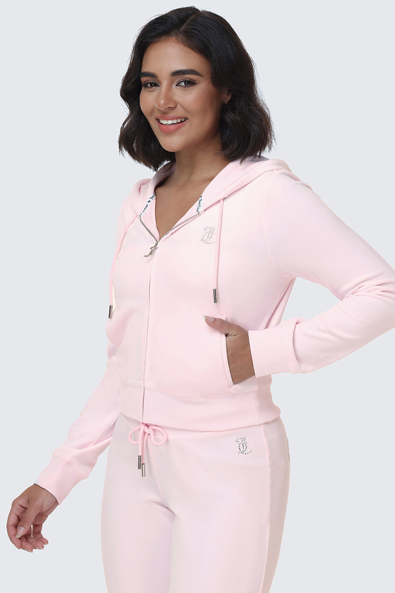 Woman smiling, wearing the OG Big Bling Velour Hoodie in light pink, styled Y2K-inspired with a subtle Juicy logo, paired with matching joggers and hands in the hoodie pockets, standing against a plain light gray background.