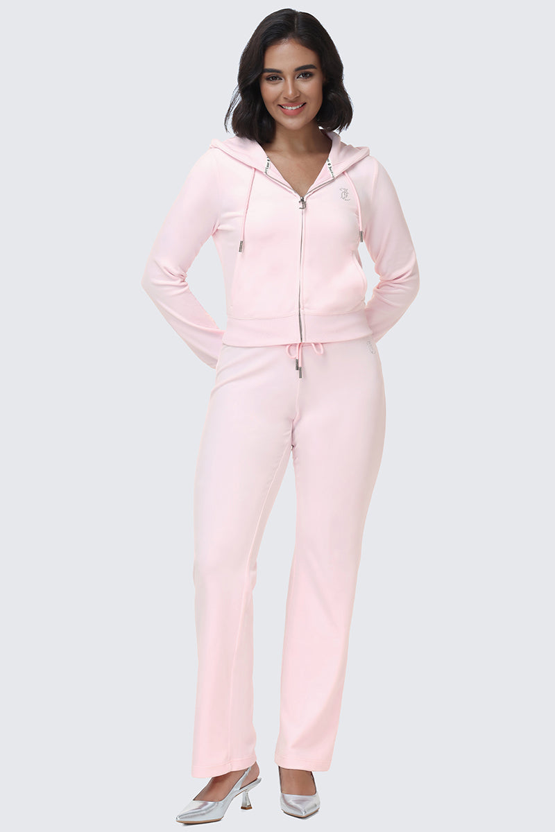 A woman stands smiling, wearing the OG Big Bling Velour Hoodie in light pink with matching drawstring pants and silver heels, against a plain light gray background.