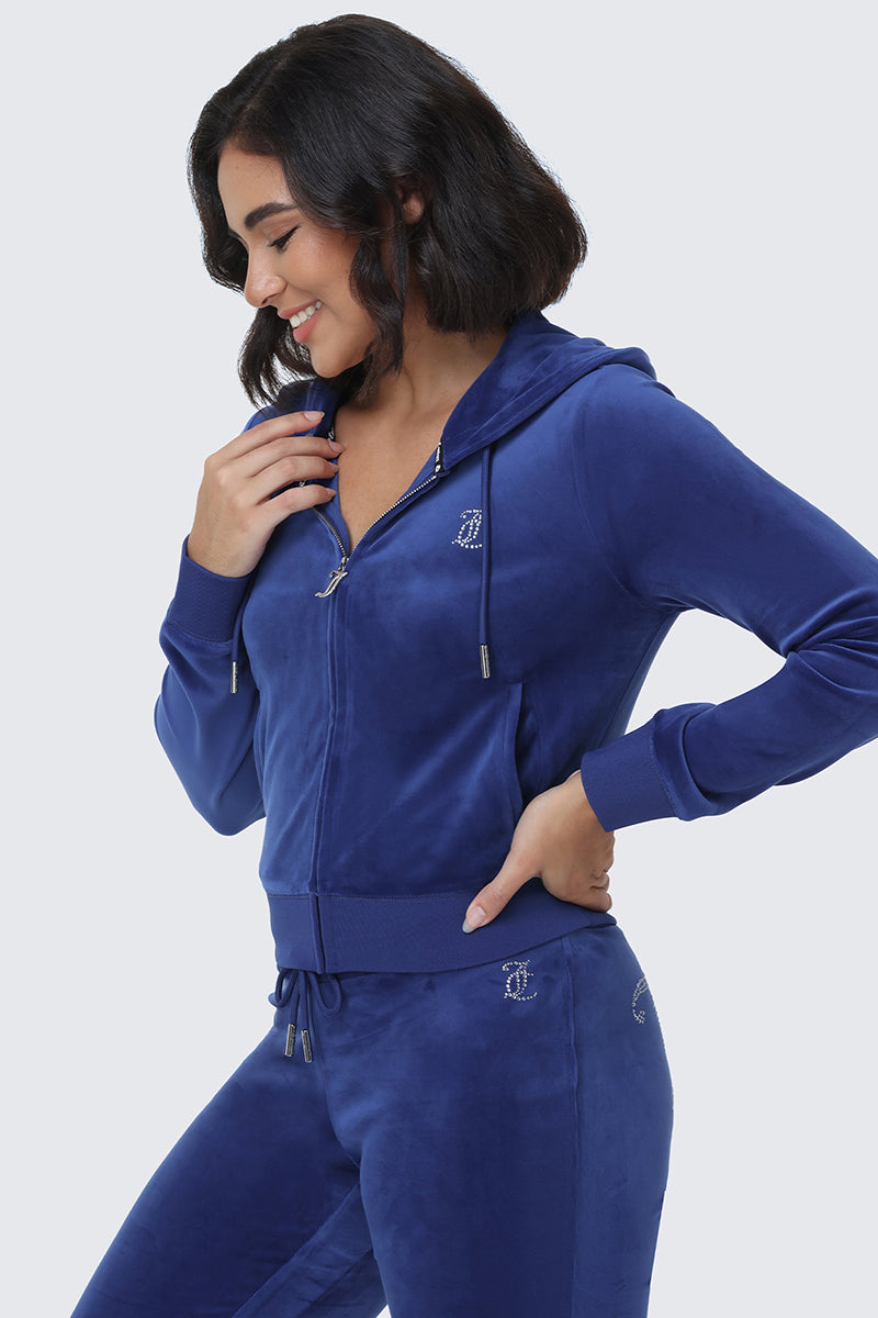 A woman smiles and looks down, wearing the OG Big Bling Velour Hoodie—a matching royal blue zip-up bedazzled hoodie paired with drawstring pants featuring a rhinestone Juicy logo—set against a light grey background for the ultimate Y2K-inspired look.