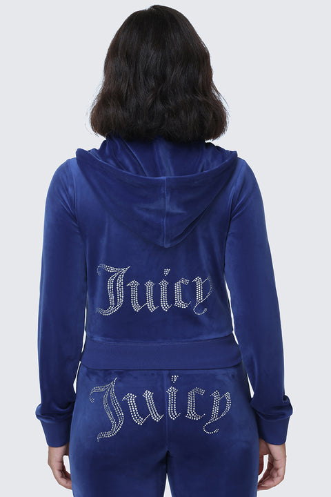 A person is seen from behind wearing the OG Big Bling Velour Hoodie, a blue velour tracksuit piece that channels iconic Y2K style. The hoodie and matching pants are both embellished with the sparkling rhinestone Juicy logo on the back.