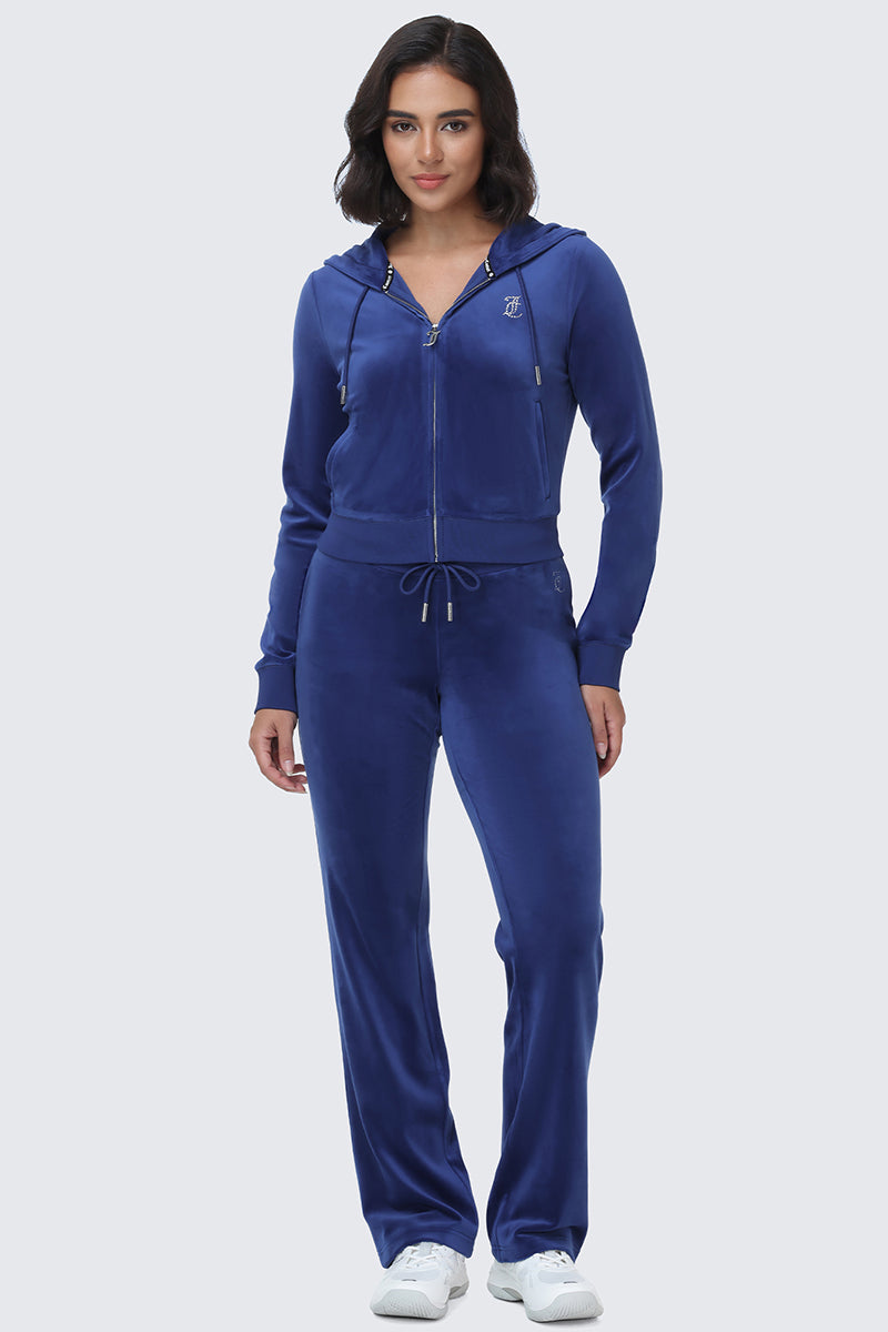 A woman stands against a plain light grey background wearing the OG Big Bling Velour Hoodie in royal blue—a zip-up velour hoodie featuring a bedazzled hood—paired with matching drawstring pants and white sneakers.