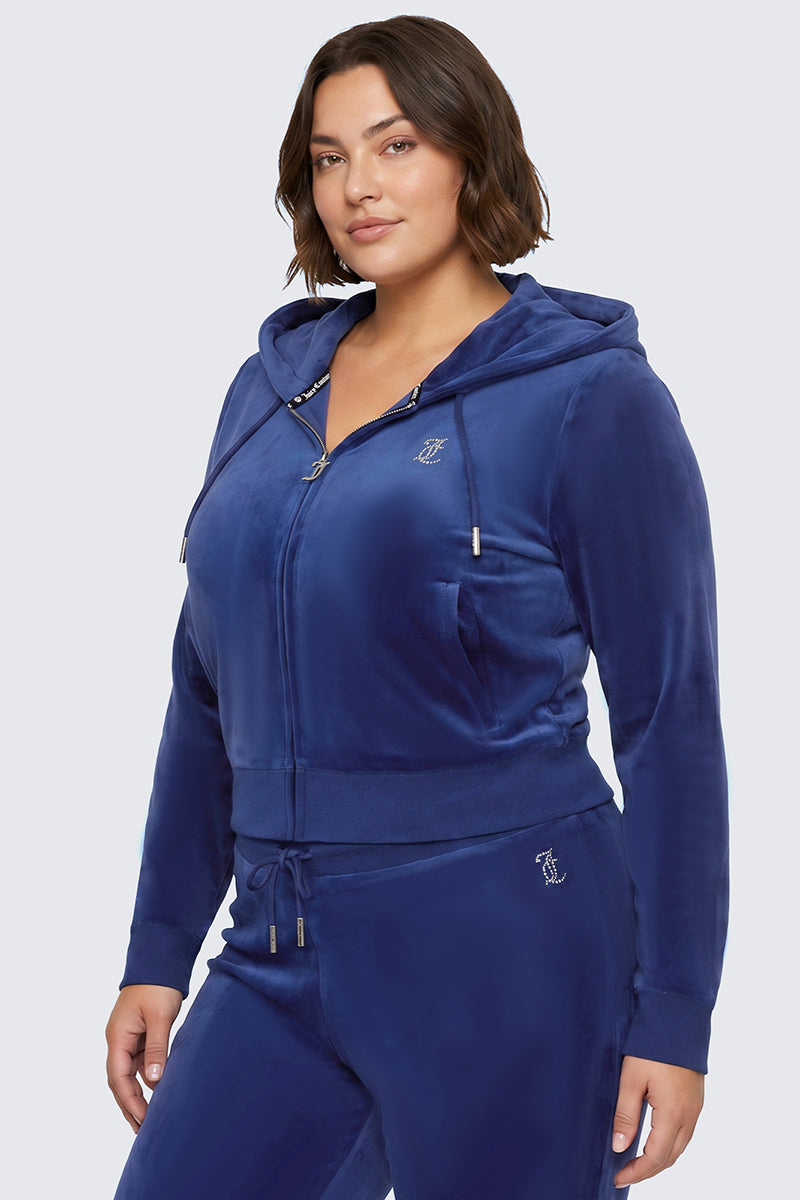 A woman is wearing the Plus-Size OG Big Bling Velour Hoodie in royal blue, styled with coordinating drawstring pants. The hoodie features a bedazzled design and a rhinestone Juicy logo, and she stands against a plain light background.