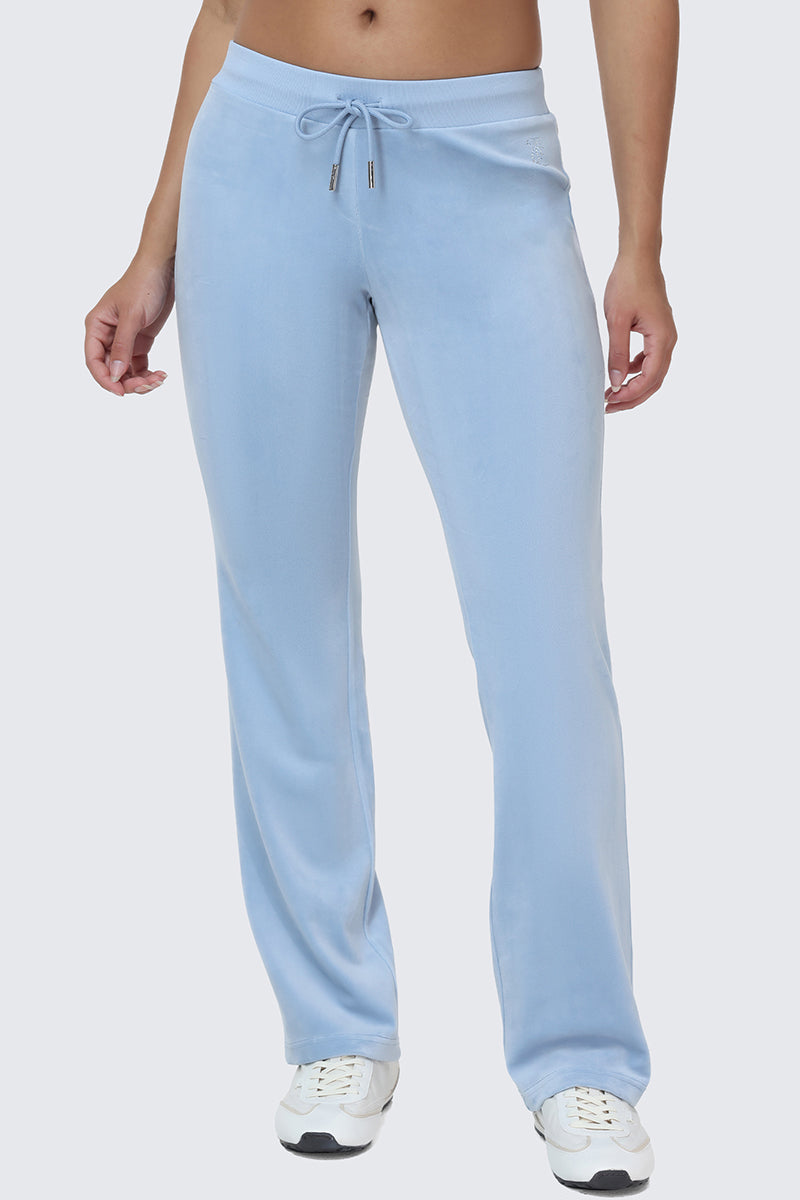 Person wearing OG Big Bling Velour Track Pants in light blue with subtle Juicy embellishment and white sneakers, standing against a plain light gray background. Upper torso and face are not visible.
