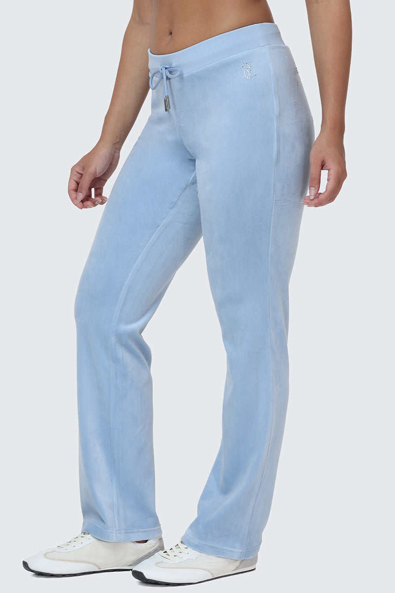A person stands against a plain background, showcasing the OG Big Bling Velour Track Pants in light blue with a subtle Y2K vibe and soft texture, paired with white sneakers.
