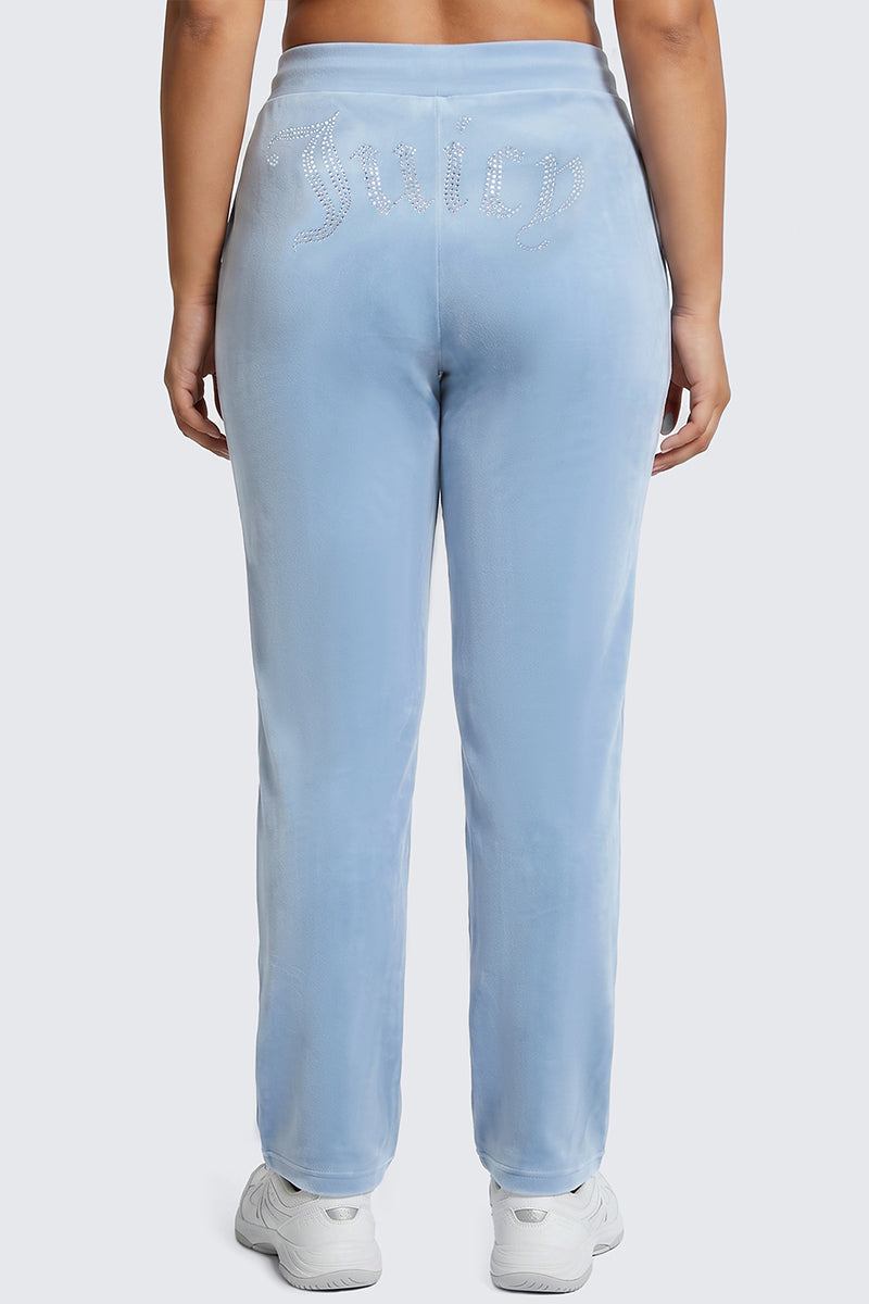 A person wearing the Plus-Size OG Big Bling Velour Track Pants in light blue, featuring a sparkling Juicy rhinestone embellishment on the back, stands facing away from the camera and channels iconic Y2K style.