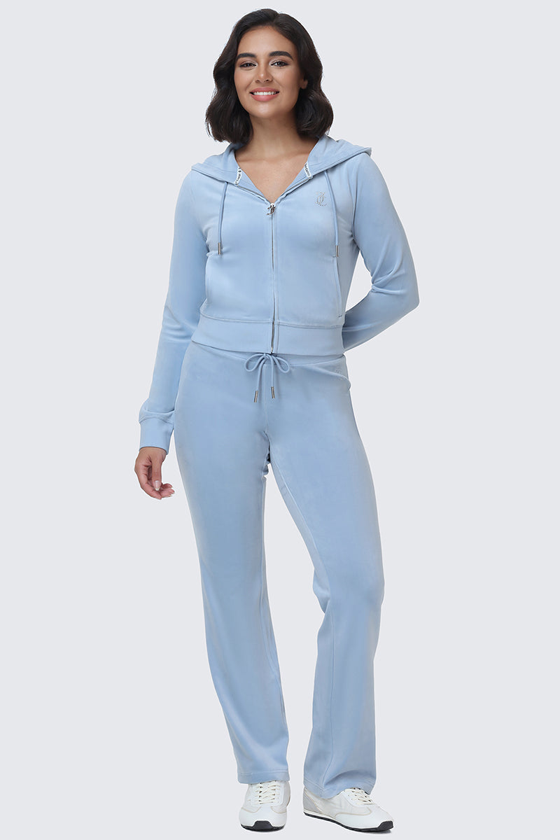 A woman stands smiling, wearing a light blue zip-up hoodie and the OG Big Bling Velour Track Pants with white sneakers, against a plain light background.