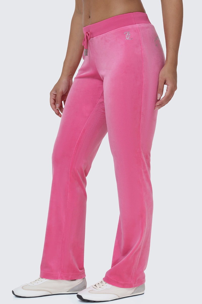 A person wearing OG Big Bling Velour Track Pants in bright pink, featuring a drawstring waist, paired with white sneakers and standing against a plain light background.
