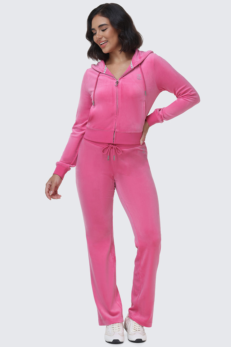 A woman smiles while wearing the OG Big Bling Velour Track Pants in bright pink with a matching zip-up hoodie and white sneakers—a playful nod to iconic Y2K style.