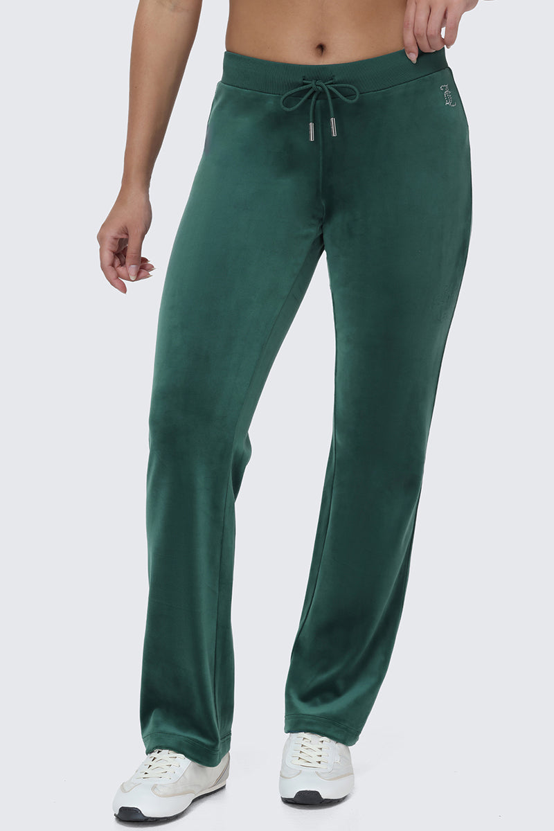 A person wearing the OG Big Bling Velour Track Pants in green, featuring subtle Juicy embellishments and styled with white sneakers, stands against a plain light gray background, channeling a laid-back Y2K vibe.