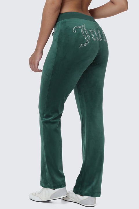 Wearing the OG Big Bling Velour Track Pants in a Y2K-inspired green with a rhinestone Juicy embellishment on the back, styled with white sneakers and standing against a plain light background.